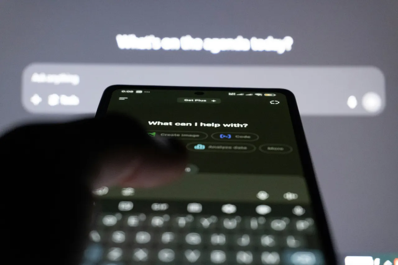 Smartphone met de ChatGPT-app en de vraag ‘What can I help with?’; op de achtergrond een beeldscherm met ChatGPT